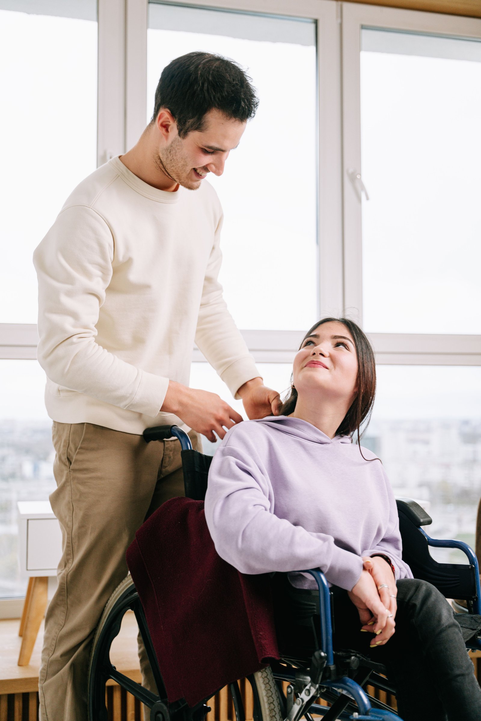 A support worker standing with a woman in a wheelchair, chatting warmly by a window.