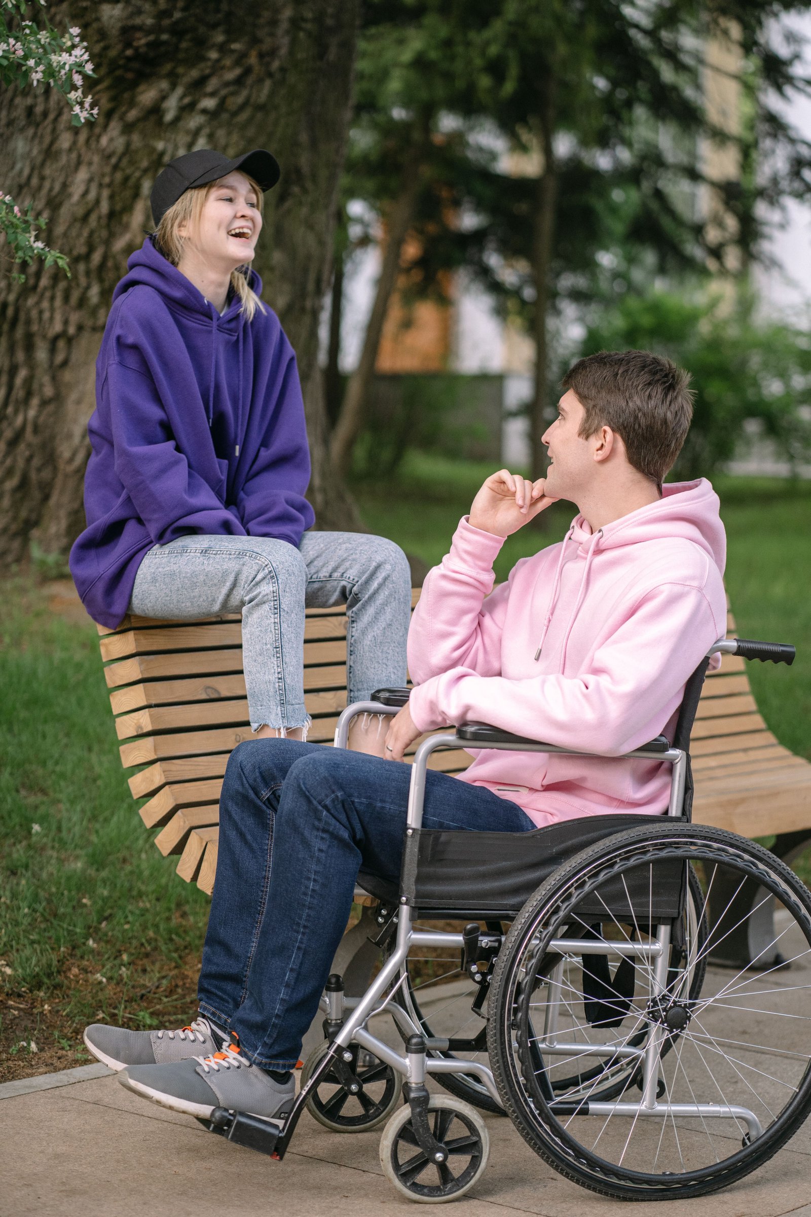 Two friends laughing together outdoors on a park bench, one of them using a wheelchair.
